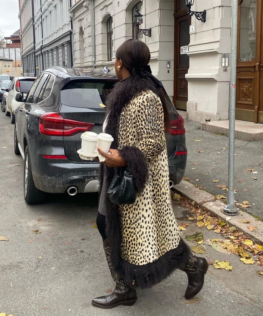 Mulher vestindo um casaco de estampa de leopardo com gola e punhos de pele preta, botas de couro marrons e segurando copos de café em uma rua urbana. Look sofisticado e ousado, combinando tendências de estampa animal e acessórios clássicos.