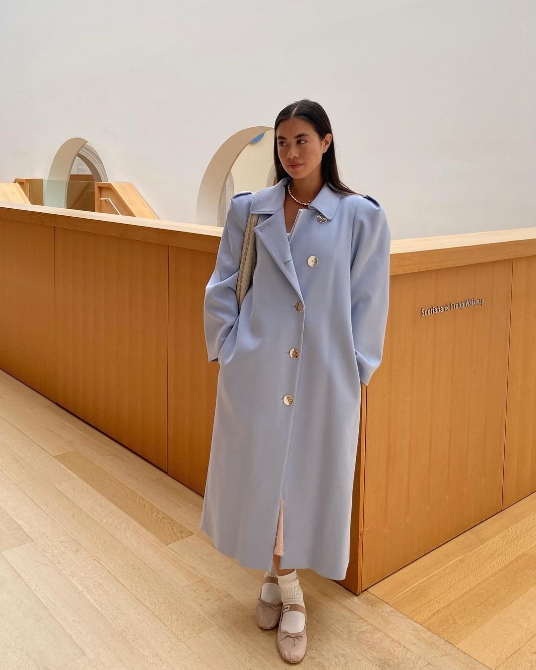 Mulher usando sobretudo oversized azul claro com ombros estruturados, combinando com sapatilhas bege e meia branca. O look minimalista destaca a tendência de peças amplas e confortáveis, ressaltando sofisticação e elegância.