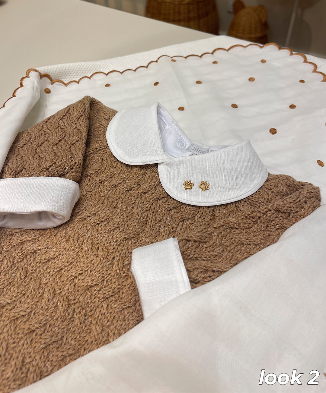 Conjunto de moda infantil em tom marrom, destacando um suéter de malha com detalhes trançados e gola em algodão branco com delicada estampa bordada. O lençol ao fundo possui bordas onduladas e pequenos pontos bordados, combinando elegância e conforto.