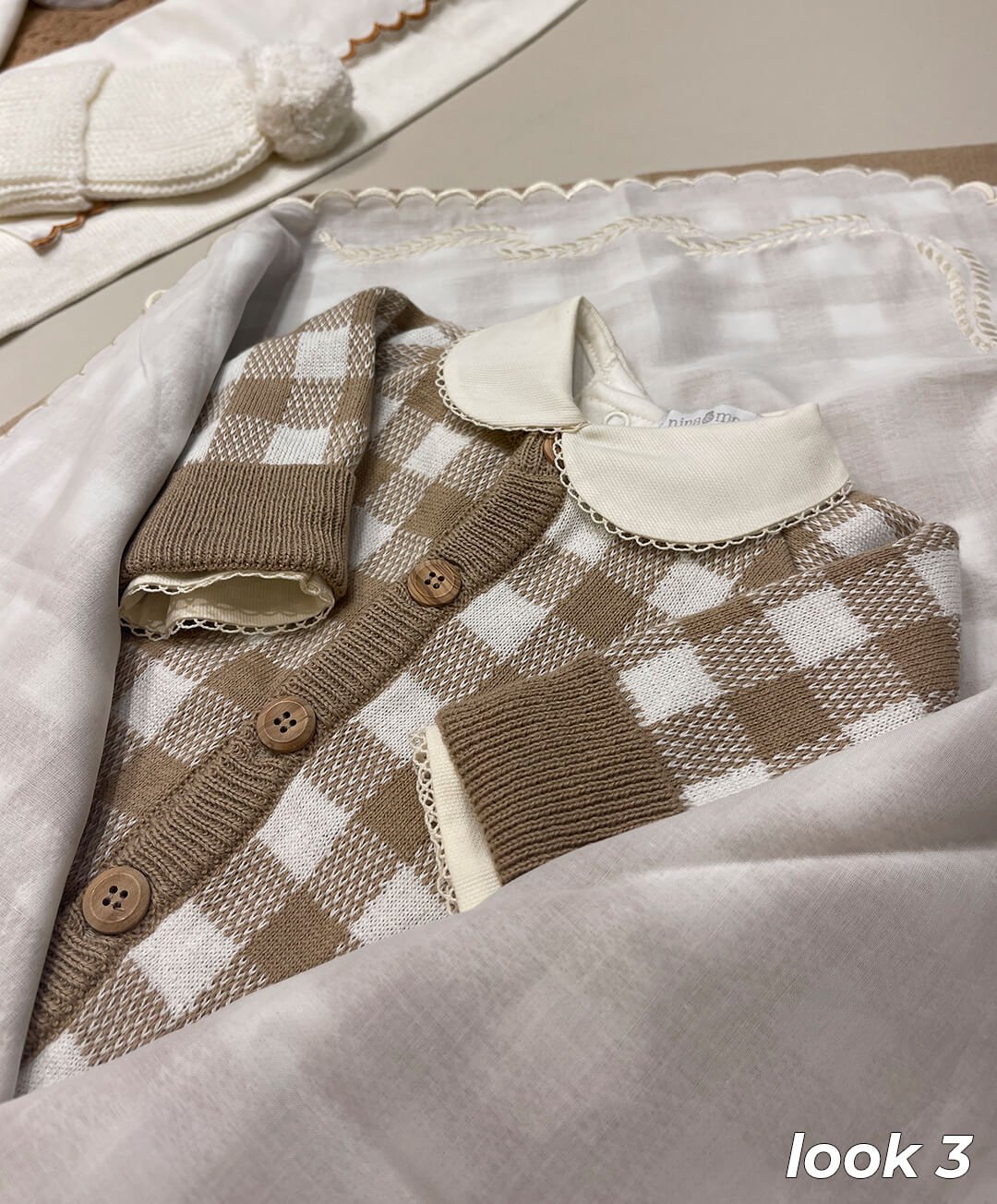 Roupa infantil estilo clássico com suéter de tricô xadrez marrom e branco. Detalhe de gola Peter Pan branca com bordado delicado. Botões de madeira e punhos canelados. Moda aconchegante e sofisticada para bebês, perfeita para o inverno.