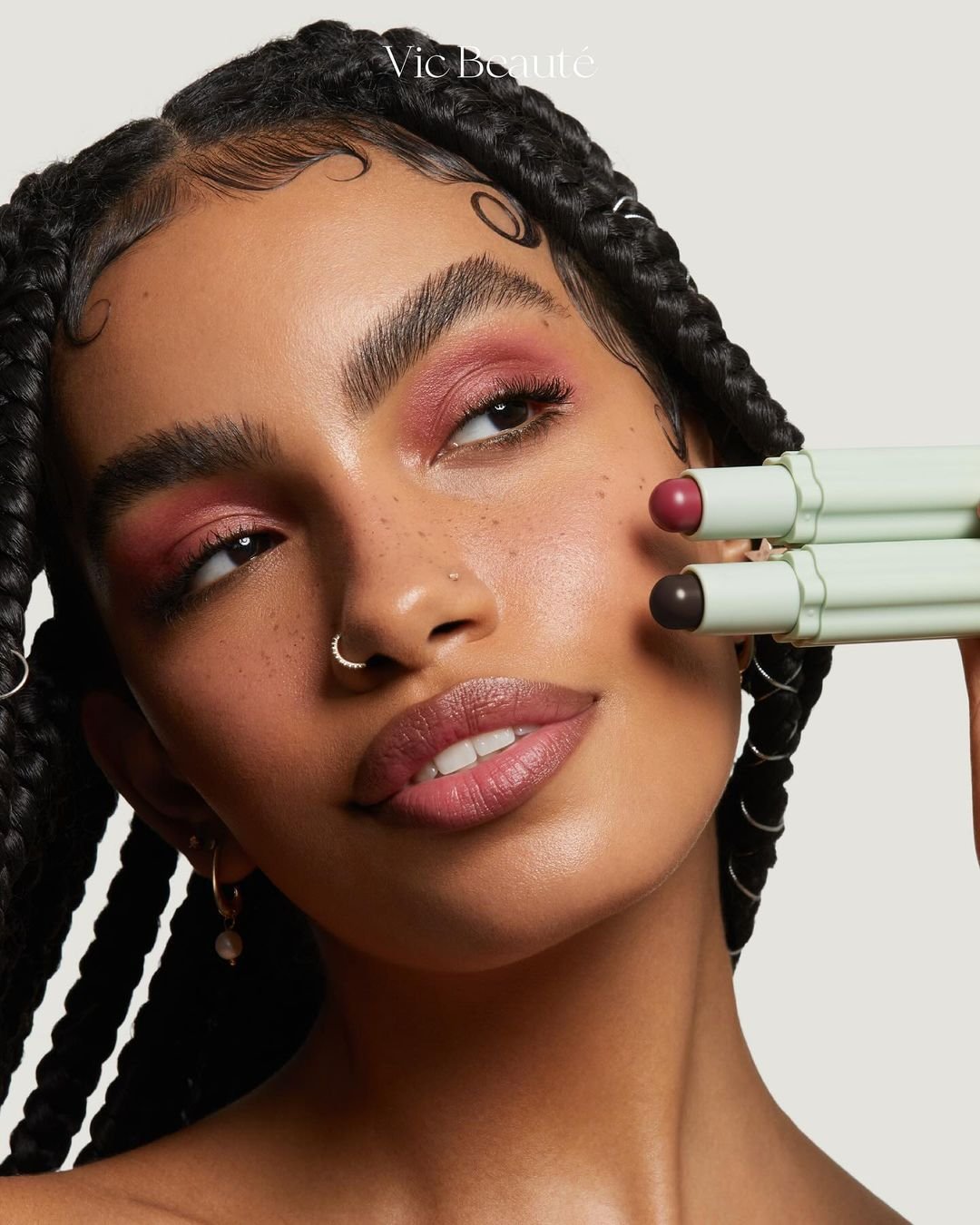 Modelo usando maquiagem vibrante com sombra rosa e batom nude. Estilo contemporâneo com tranças box braids longas e ajustadas. Piercings no nariz e brincos de argola. Representa tendências modernas de beleza e autoexpressão.