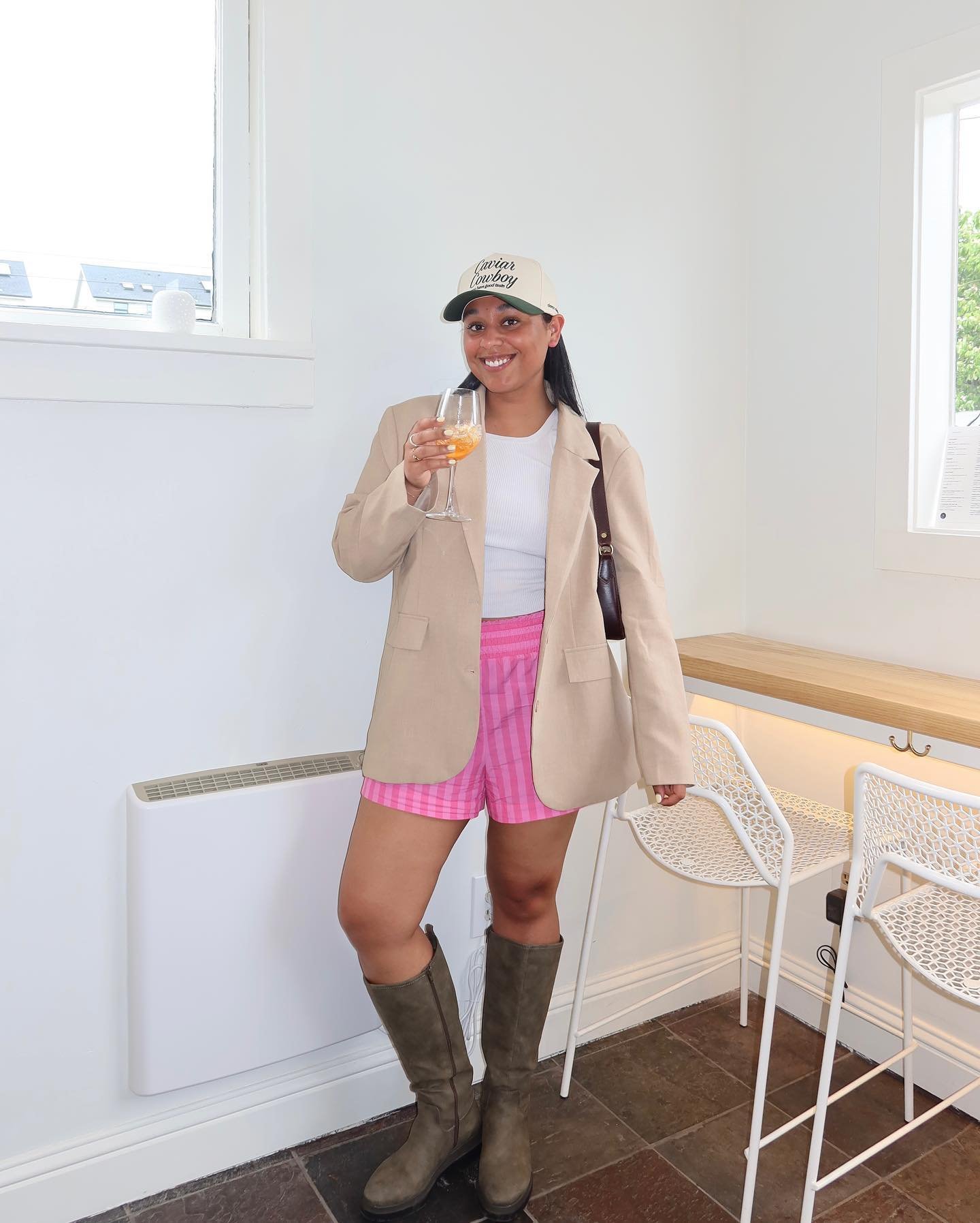 Mulher veste blazer bege, camiseta branca, shorts xadrez rosa e botas de cano longo cinza. Completa o look com boné bege e bolsa lateral marrom, segurando taça de bebida. Mistura estilo casual com elementos clássicos.