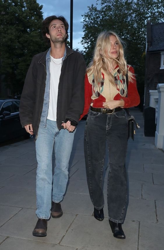 Casal caminhando na rua. Ele veste jaqueta escura, camiseta branca, jeans claros e botas marrons. Ela usa blusa vermelho e bege, jeans escuros de cintura alta, cinto preto e botas pretas. Estilo casual e retrô, influenciado por tendências dos anos 90.