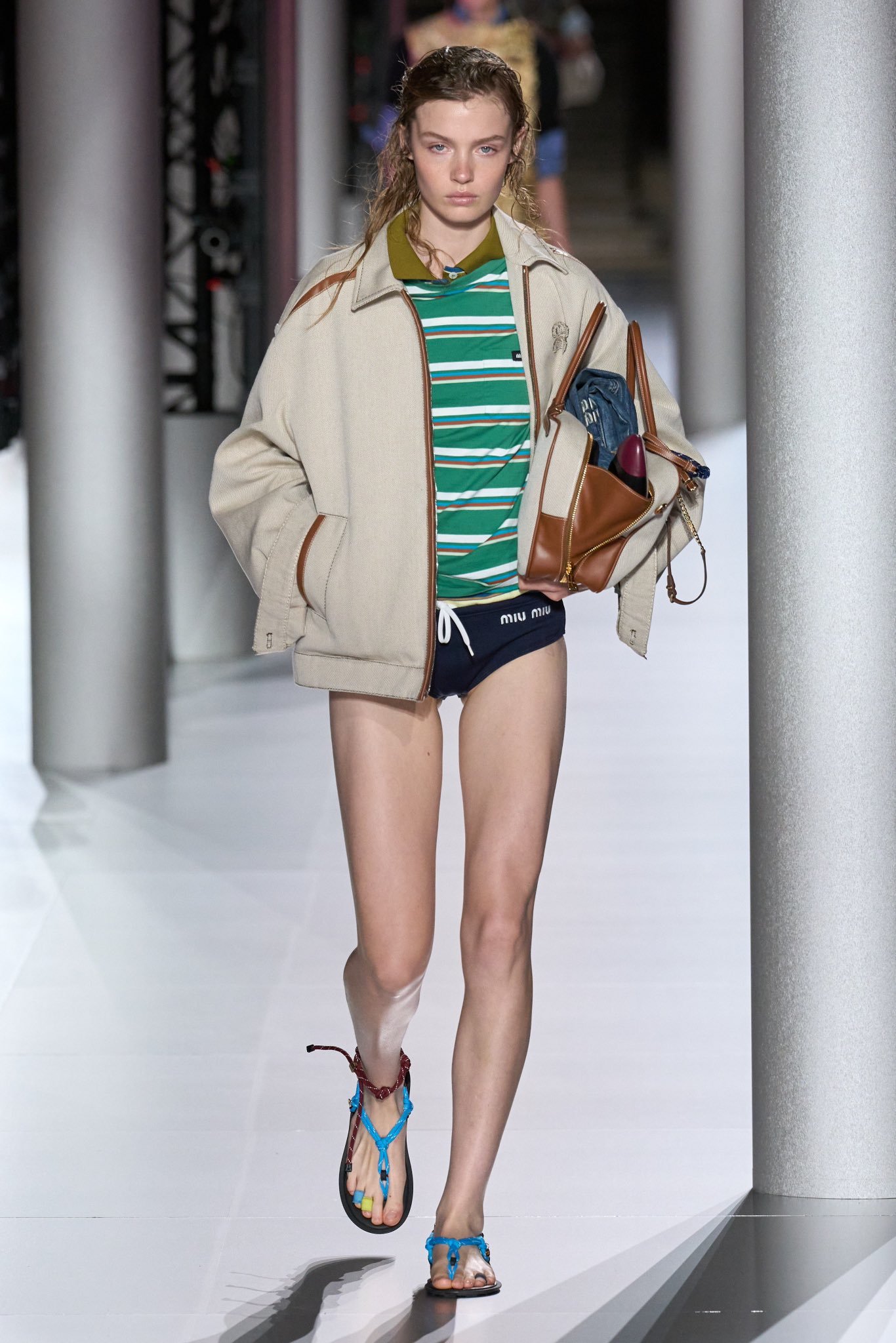 Modelo desfilando com blusa polo listrada, jaqueta bege oversized, e shorts esportivo da Miu Miu. Look complementado por bolsa de couro e sandálias minimalistas em cores vivas. Combinação de estilo casual e sofisticado.