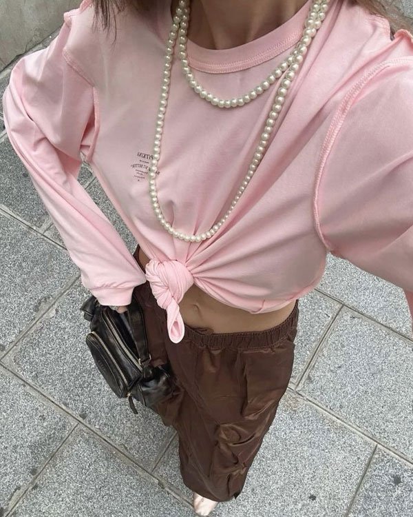 Look casual e moderno com blusa rosa clara, amarrada na cintura, combinada com calça cargo marrom. Colar de pérolas longo como acessório, dando um toque vintage ao visual. Ideal para indexar em moda jovem e tendências urbanas.