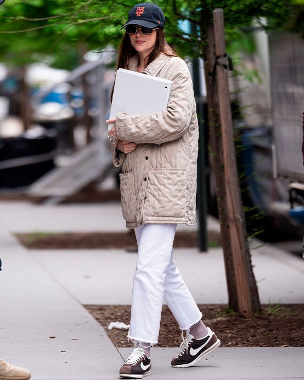 Pessoa usando look casual com tendências modernas. Ela veste um casaco acolchoado bege oversized, calça branca cropped e tênis Nike preto e branco. Acessórios incluem boné preto e óculos de sol. Carrega um laptop, estilo urbano e confortável.