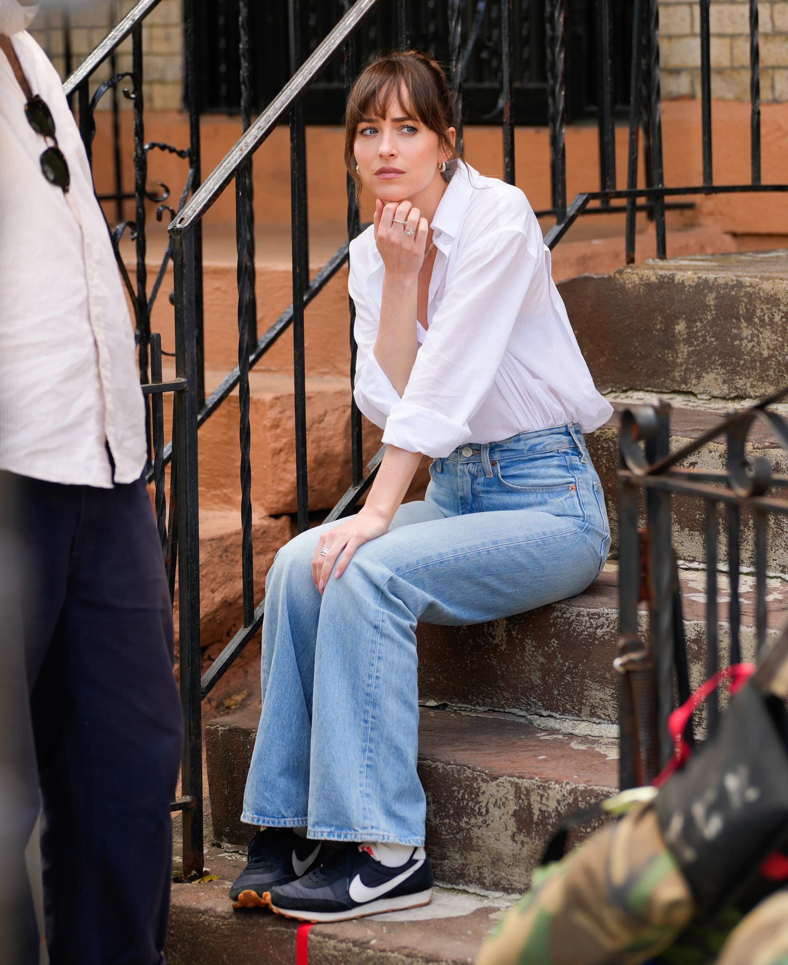 Mulher vestindo camisa branca com mangas dobradas, calça jeans de cintura alta e tênis Nike pretos. O visual é casual e descontraído, combinando peças clássicas e confortáveis, tendência popular para um estilo urbano e prático.