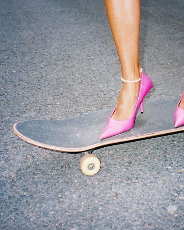 Mulher andando de skate usando salto alto rosa e tornozeleira de pérolas brancas, combinando moda ousada e urbana. Tendência 