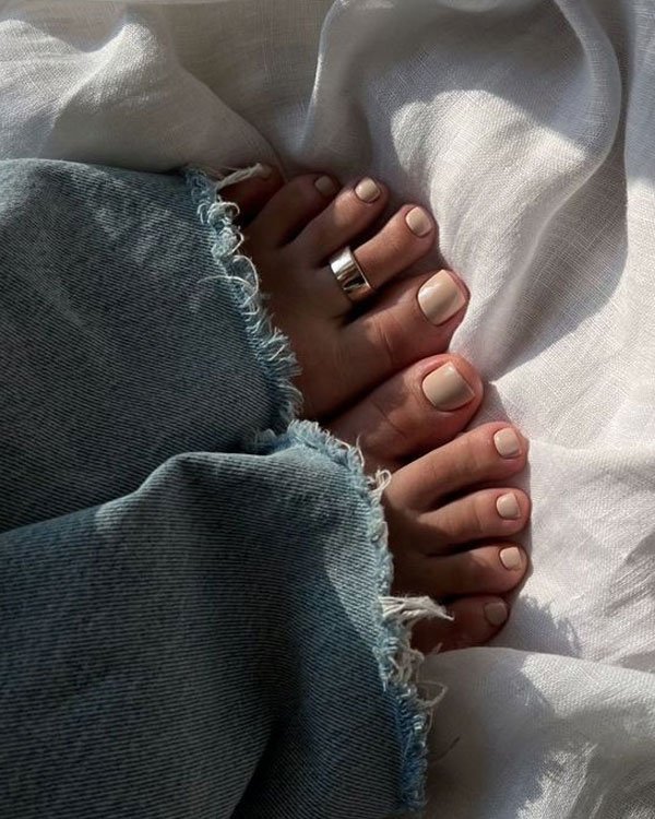 Jeans largos com bainha desfiada, tendência casual e moderna. Unhas dos pés pintadas de nude, combinando com um anel prateado. Look despojado que destaca detalhes minimalistas e confortáveis na moda atual.