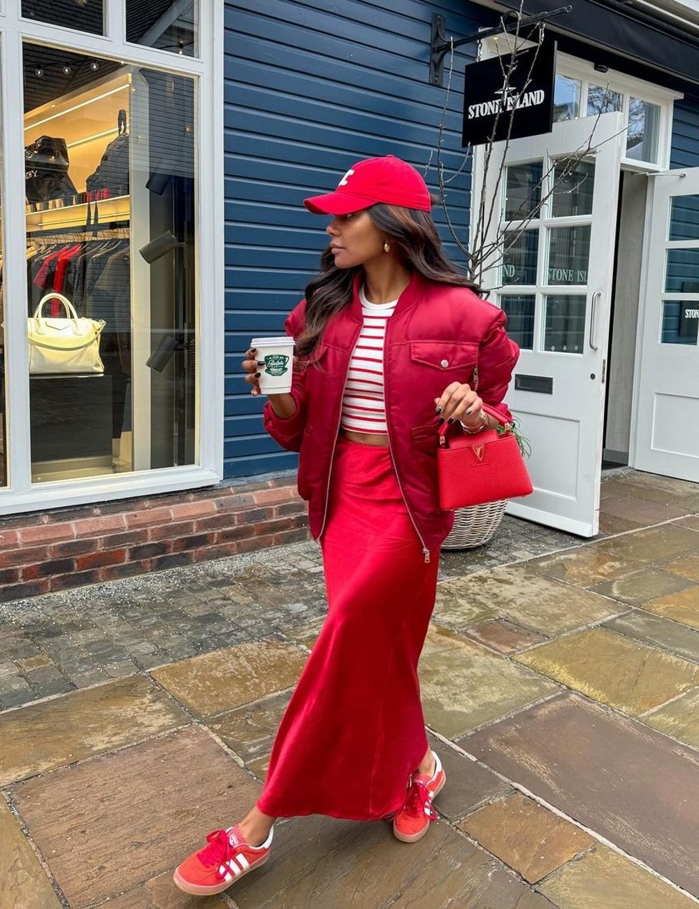 Mulher com look fashion. Usa boné vermelho, jaqueta bomber, saia longa vermelho-escarlate, camiseta listrada e tênis vermelho. Acessórios incluem uma bolsa de mão vermelha e um copo de café. O que está em alta.