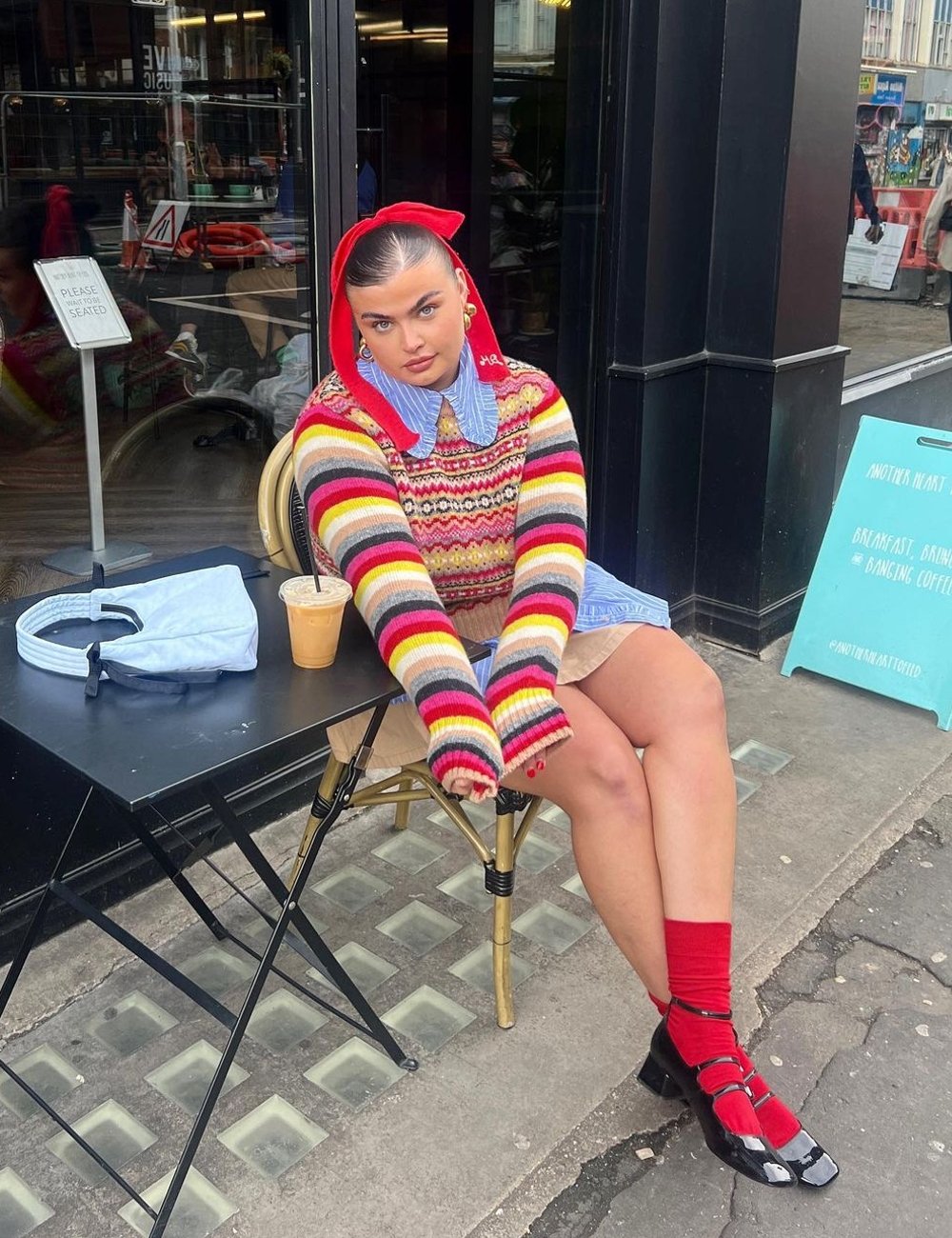 Pessoa sentada em café usando suéter colorido listrado, sobre camisa azul de gola e chapéu vermelho. Calça meias vermelhas combinando com a faixa da cabeça e sapatos pretos de salto baixo. O que está em alta.