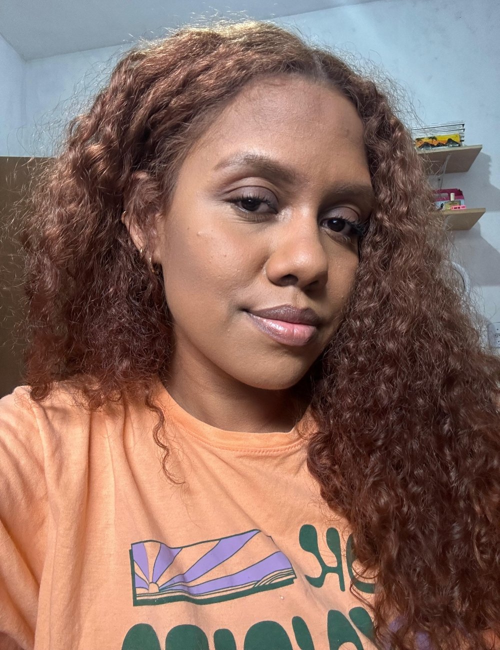 Mulher com cabelo cacheado castanho claro, utilizando uma camiseta casual laranja com uma estampa gráfica verde e roxa. A maquiagem é discreta, usando paleta multifuncional. Ambiente interno com prateleiras ao fundo.