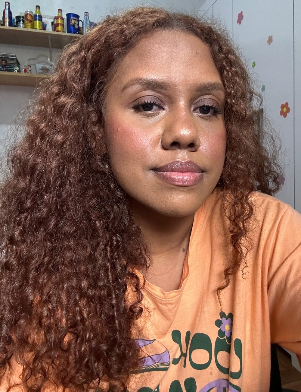 Mulher com cabelo cacheado solto, vestindo camiseta laranja com estampa floral e de letras verdes. Visual casual e descontraído, seguindo tendência de conforto e estilo jovem. Maquiagem com paleta multifuncional, destacando o brilho natural da pele.