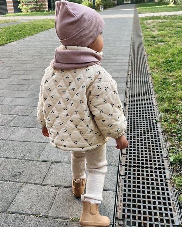 Criança com look de inverno, vestindo jaqueta acolchoada estampa floral, gorro e cachecol rosa, calça térmica branca e botas bege. Estilo aconchegante e tendência de moda infantil.