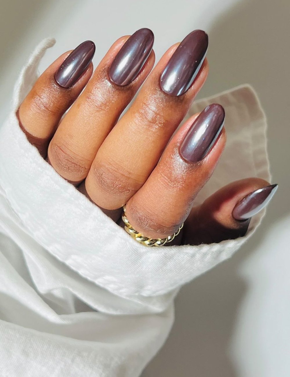 Unhas longas e bem cuidadas, pintadas com esmalte marrom e pó cromado, combinadas com anel de ouro delicado. A manga branca de camisa confere um toque elegante e minimalista, destacando tendências de moda e estilo sofisticado.