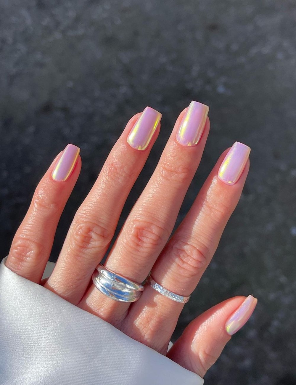 Unhas com esmalte rosa e pó cromado. Uso de anéis prateados e jaqueta branca de cetim, complementando um estilo sofisticado e contemporâneo. Ideal para indexar buscas relacionadas a moda, manicure e acessórios femininos.