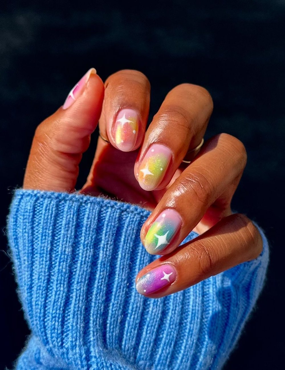 Unhas decoradas em degradê de tons pastéis, com estrelas brilhantes, com pó cromado. A mão veste um suéter azul de malha canelada, tendência outono/inverno 2023, destacando uma combinação harmoniosa entre moda e arte nas unhas.