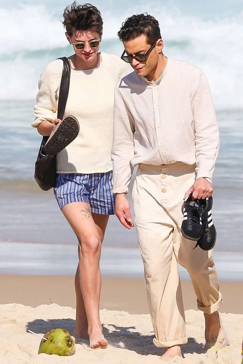 Casal passeia na praia com estilo casual. Ele veste camisa leve bege e calça clara com os punhos dobrados. Ela usa suéter creme e shorts curtos listrados azul e branco. Ambos estão descalços e usando óculos escuros, tendência despojada e confortável.