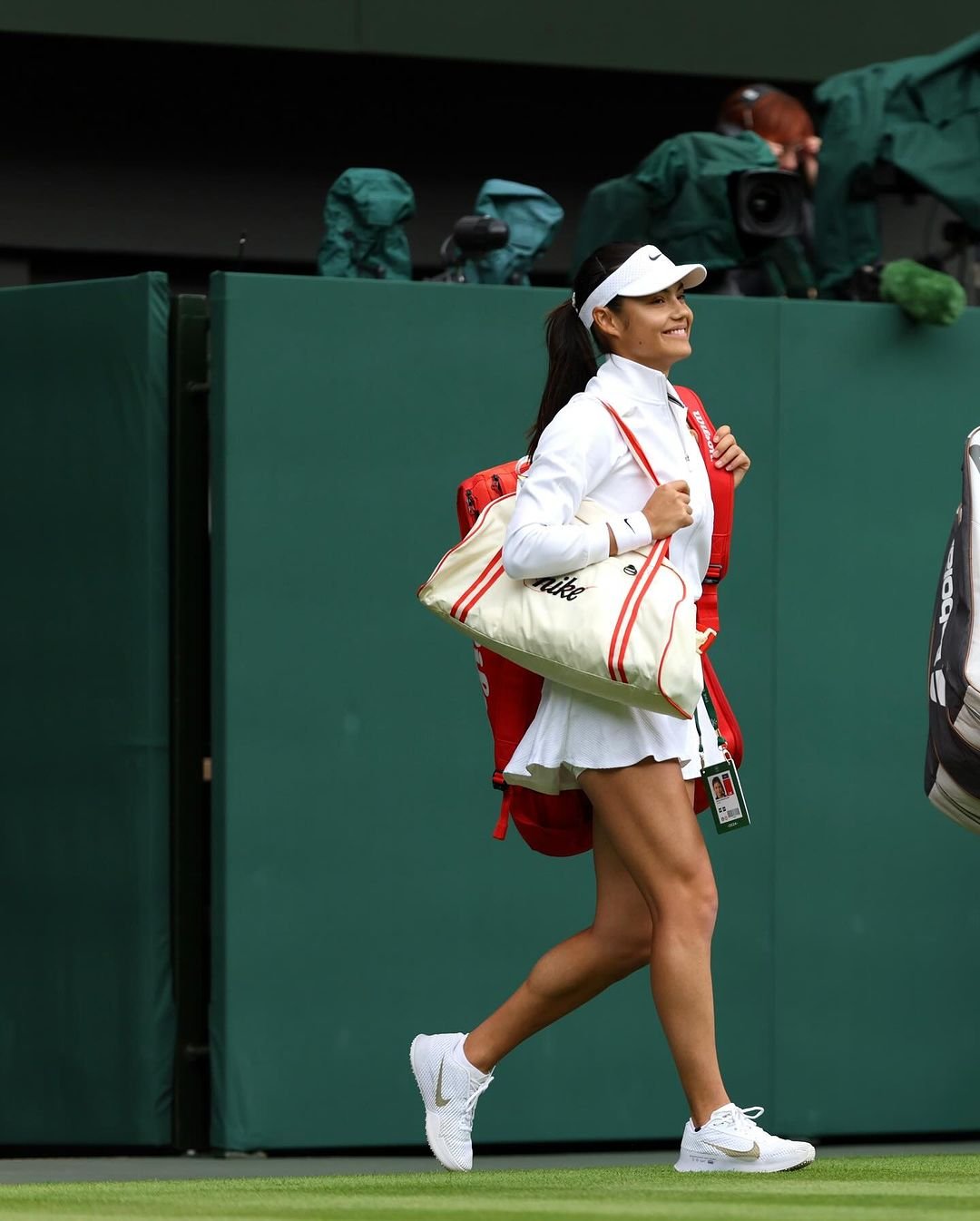 Tenista usando trajes esportivos brancos da Nike com detalhes vermelhos, incluindo saia, camiseta e jaqueta. Completa o look com boné branco e tênis esportivos brancos. Estilo clássico e elegante para competições de tênis. produtos de beleza