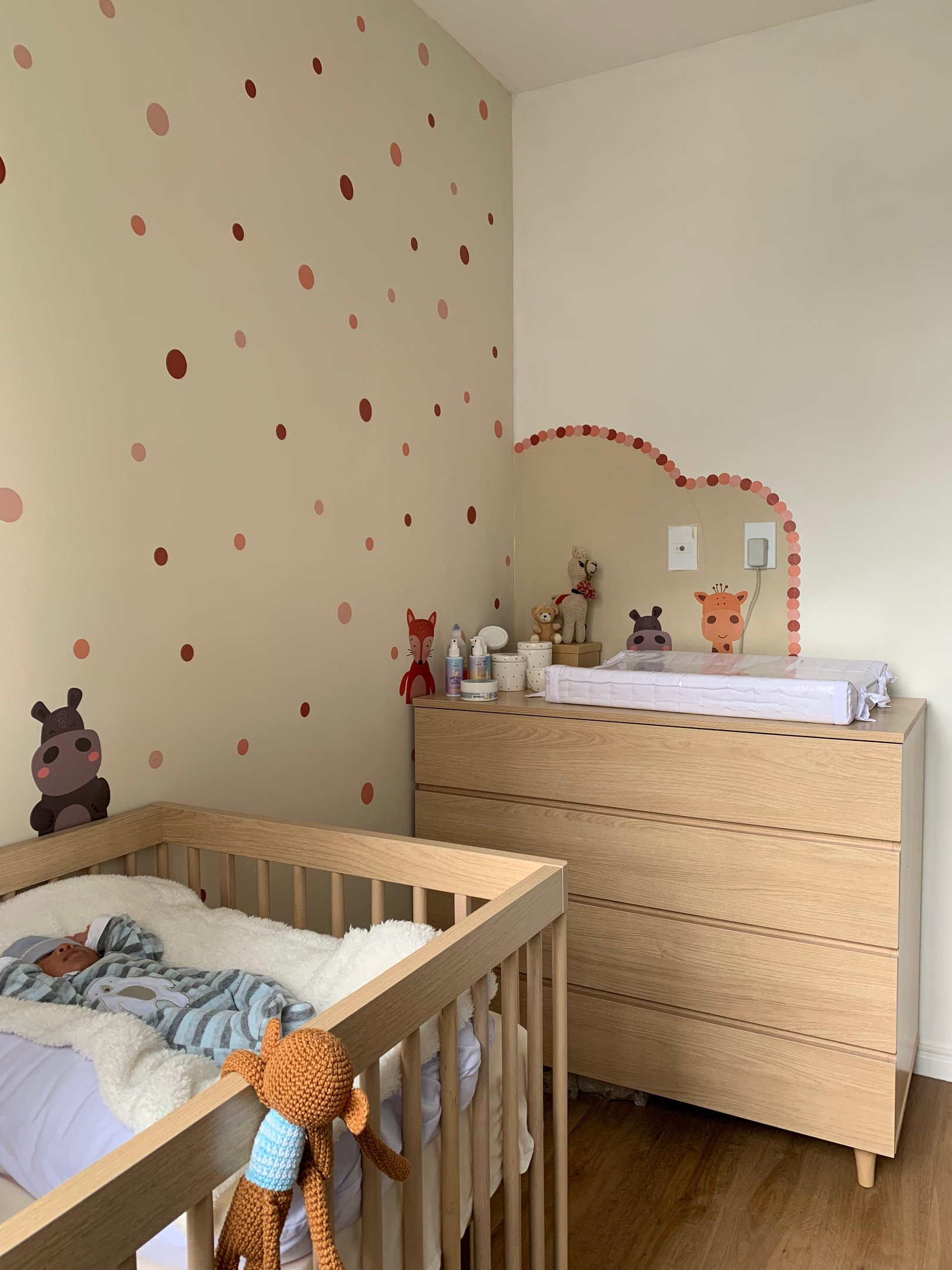 Quarto de bebê decorado com papel de parede de bolinhas, móveis de madeira clara e pelúcias. O berço possui cobertor de lã. As tendências incluem decoração minimalista e tons neutros, criando um ambiente aconchegante e moderno para o bebê.