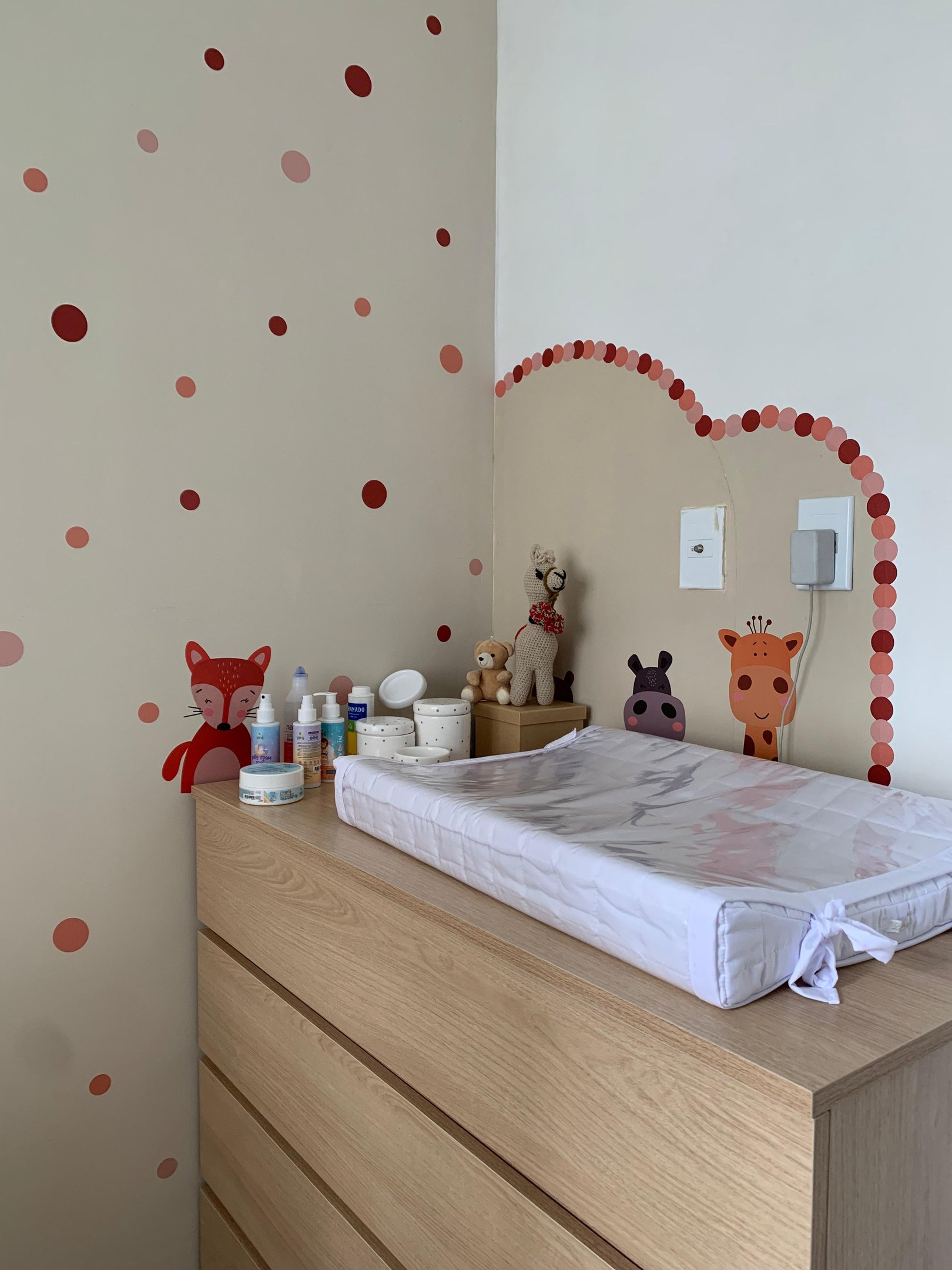 Quarto de bebê decorado com tema de animais e cores suaves. Paredes com adesivos circulares em tons de vermelho e rosa, trocador sobre cômoda de madeira clara, e pelúcias. Estilo moderno e acolhedor, ideal para quartos infantis.