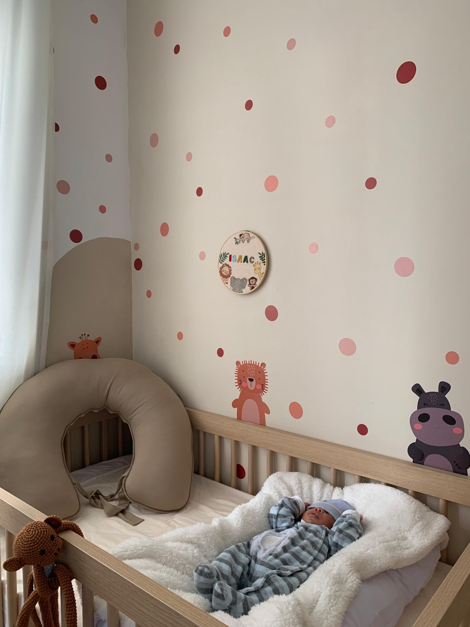 Imagem de um quarto infantil decorado com estilo moderno e minimalista, apresentando um berço de madeira clara com roupa de cama branca. O bebê está vestido com um macacão de listras azuis e brancas, tendência de moda aconchegante e confortável.