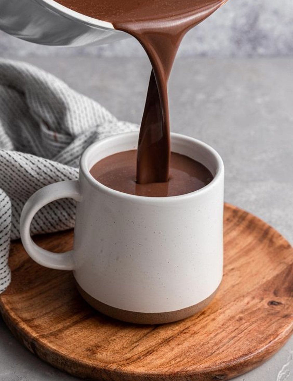 Imagem de uma bebida quente, possivelmente chocolate, sendo vertida em uma caneca branca de cerâmica sobre um suporte de madeira. Receitas para o frio.