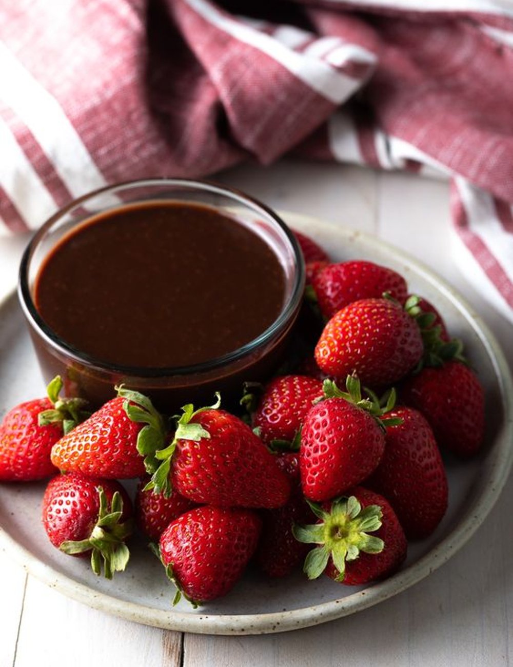 Imagem de morangos frescos ao lado de um recipiente com calda de chocolate. Ao fundo, um tecido vermelho com detalhes brancos. Receitas para o frio.