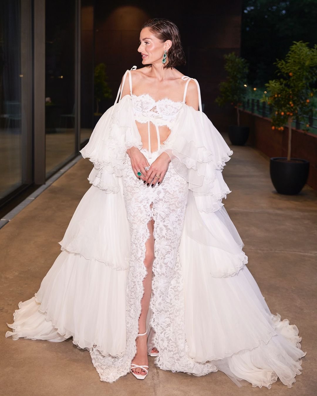 Mulher usando um elegante vestido branco de renda e tule, com mangas volumosas e detalhes recortados. O look é complementado por sandálias de salto alto e acessórios discretos, refletindo tendências contemporâneas de moda nupcial e alta-costura.