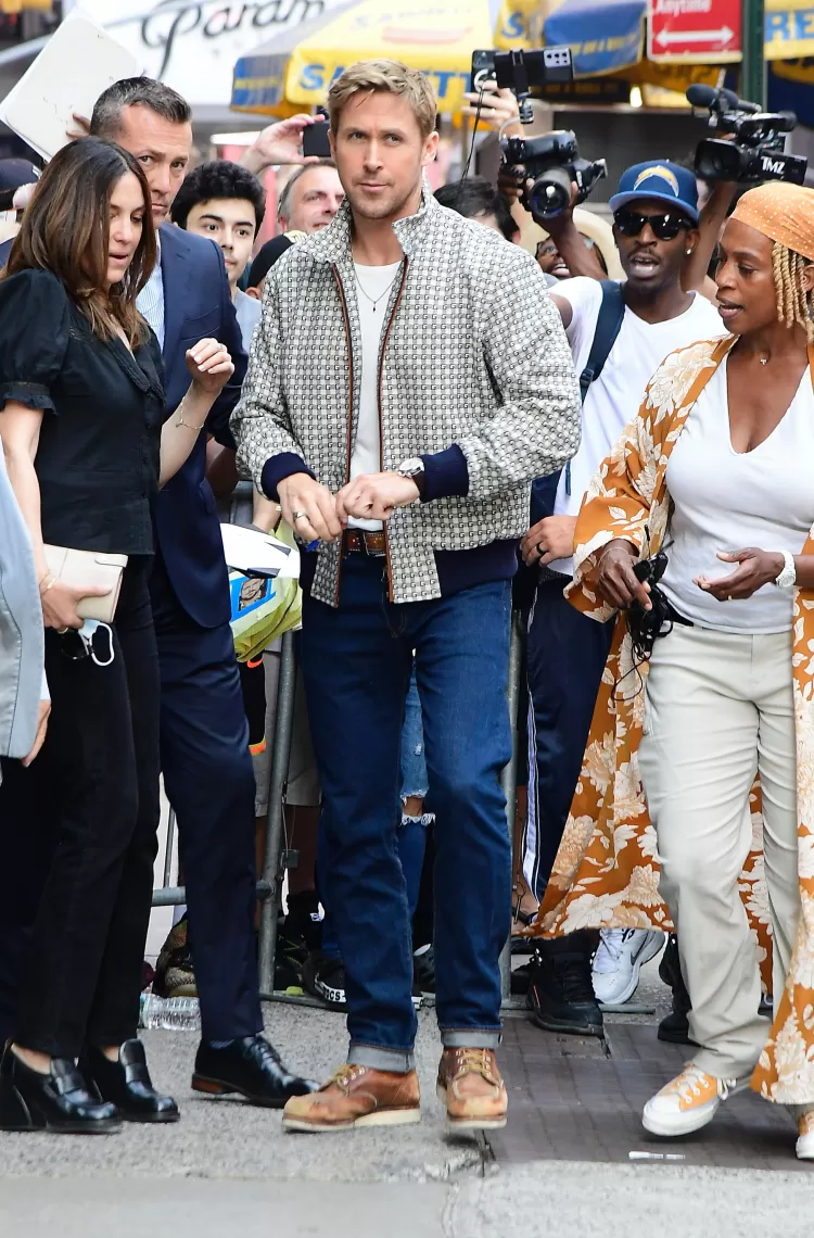 Homem vestindo jaqueta estampada, camiseta branca, jeans e mocassins. Ao lado, mulher com blusa preta e calça preta, outra mulher veste casaco floral laranja e branco, com camiseta branca e calça bege. Tendências urbanas e casuais marcando presença.