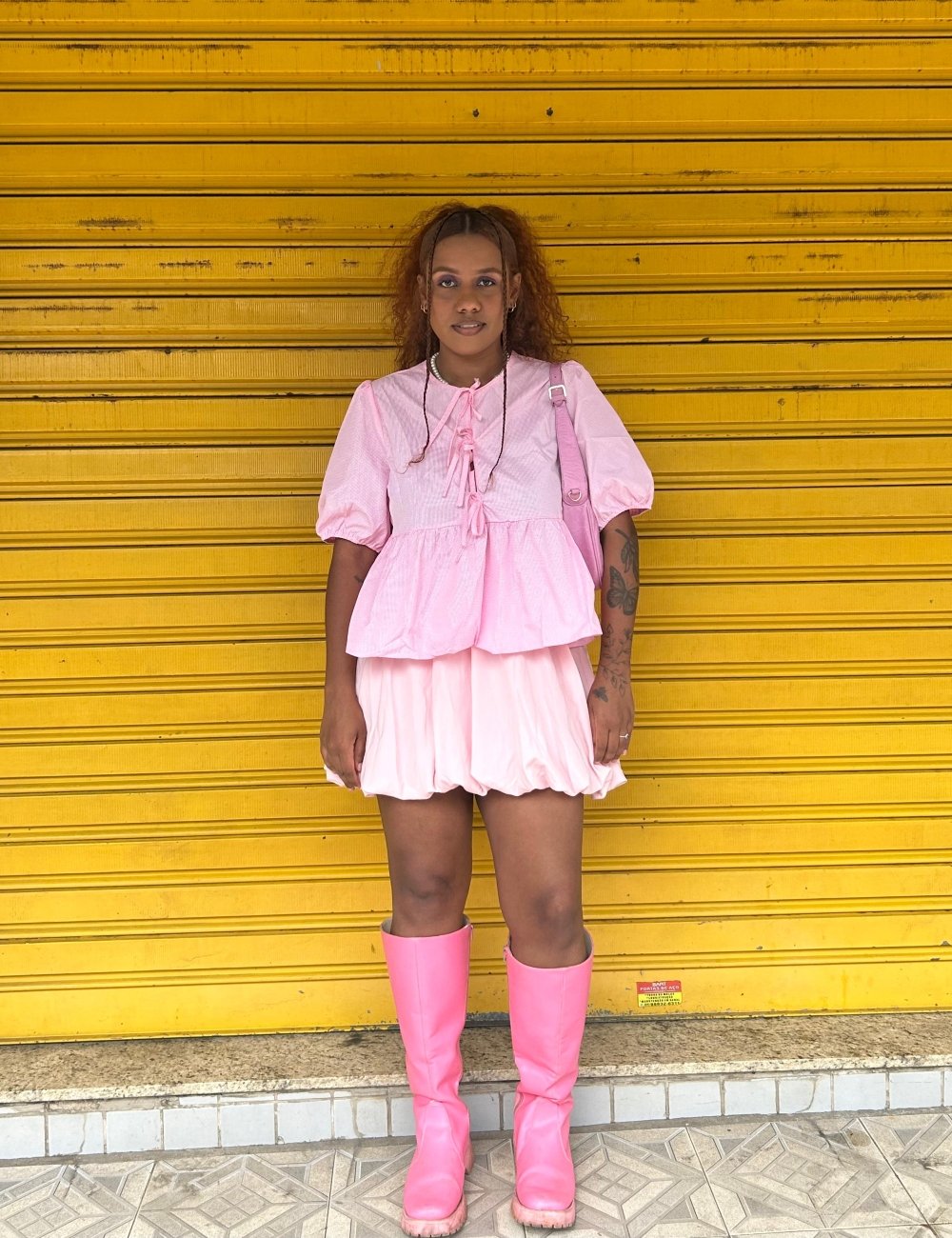 Pessoa vestida com look monocromático rosa, composto por blusa com mangas bufantes, saia balonê e botas de cano alto. A moda evidencia um estilo ousado, vibrante e feminino, com tendência de roupas com texturas e cores chamativas.