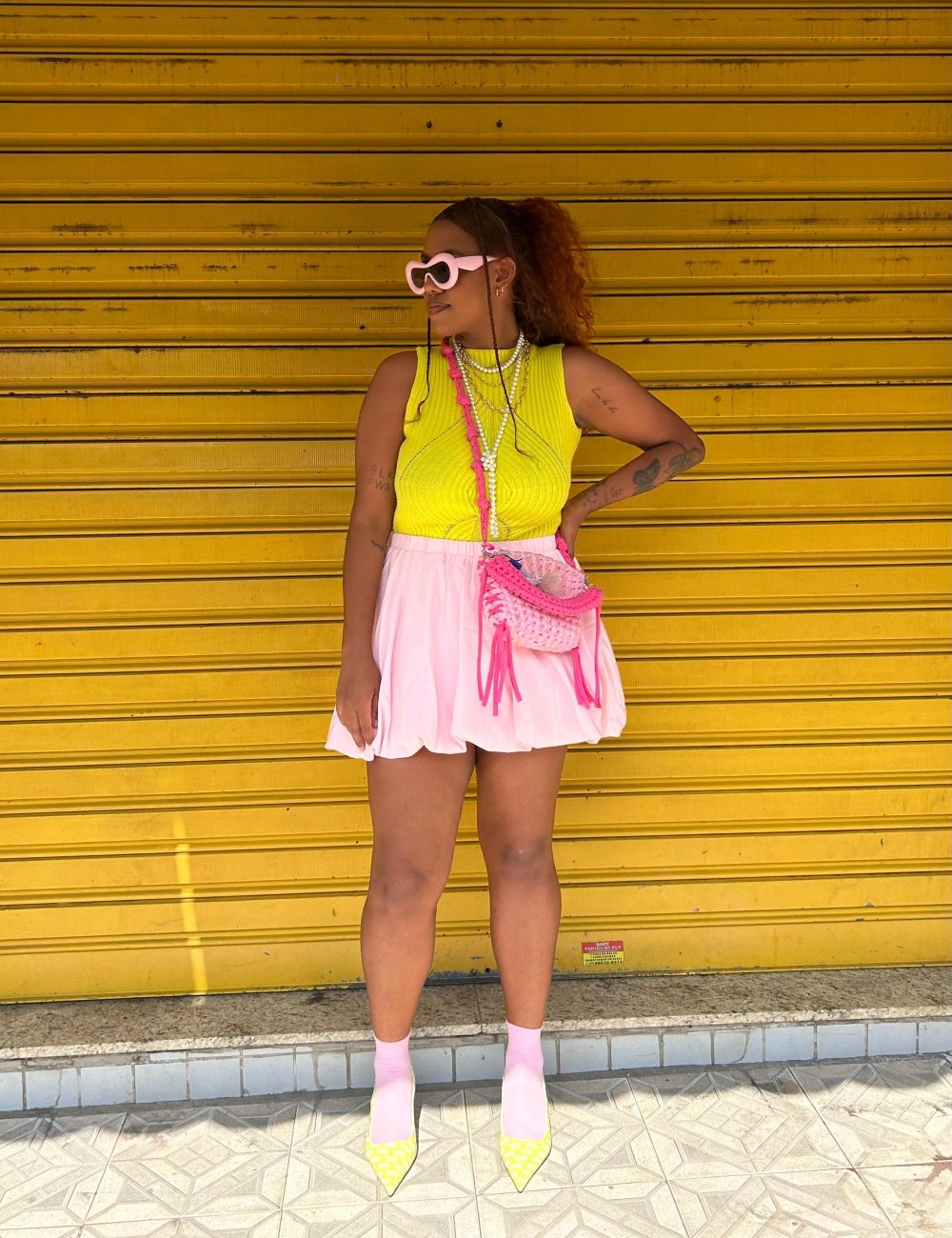 Jovem com blusa amarela sem mangas, saia balonê, óculos de sol rosa, bolsa de tricô rosa e amarelo. Complementa o look com sapatos de salto amarelos e meias cor de rosa. Estilo vibrante e moderno, destacando tendências coloridas e ousadas.