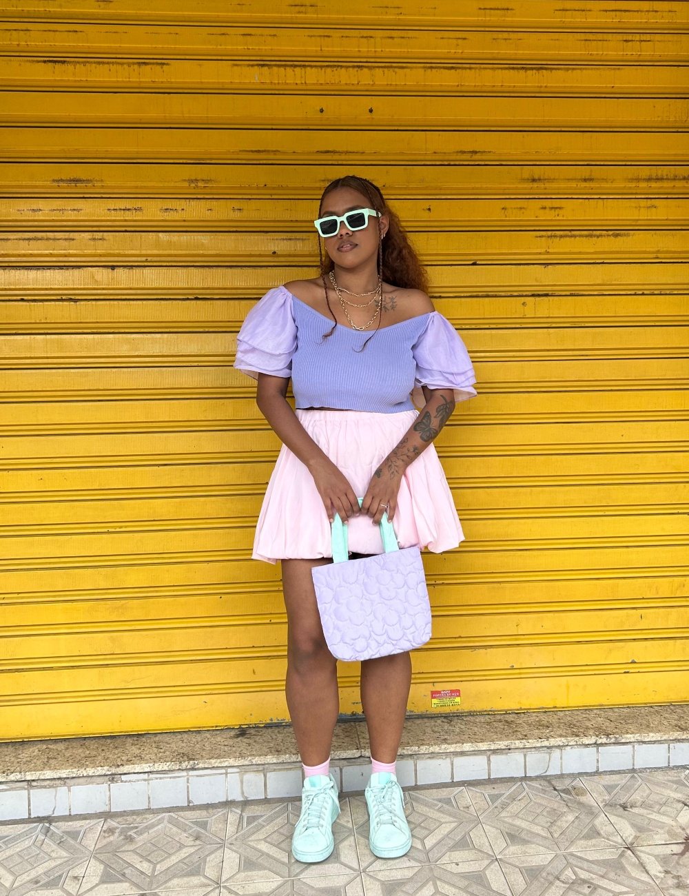 Mulher vestindo blusa lilás de mangas bufantes e saia balonê, bolsa lilás e tênis verde pastel. Acessoriza com óculos de sol branco e colares dourados. Look casual, inspirado nas tendências de moda primavera-verão com cores pastel e femininas.