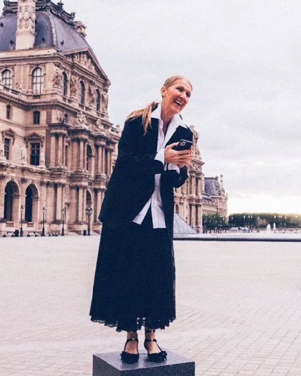 Mulher em frente ao Louvre, Paris, veste camisa branca de botões, blazer preto oversized e saia midi preta. Look une elegância clássica com tendências contemporâneas, sapatos de bico quadrado e cabelo preso em rabo de cavalo despojado.