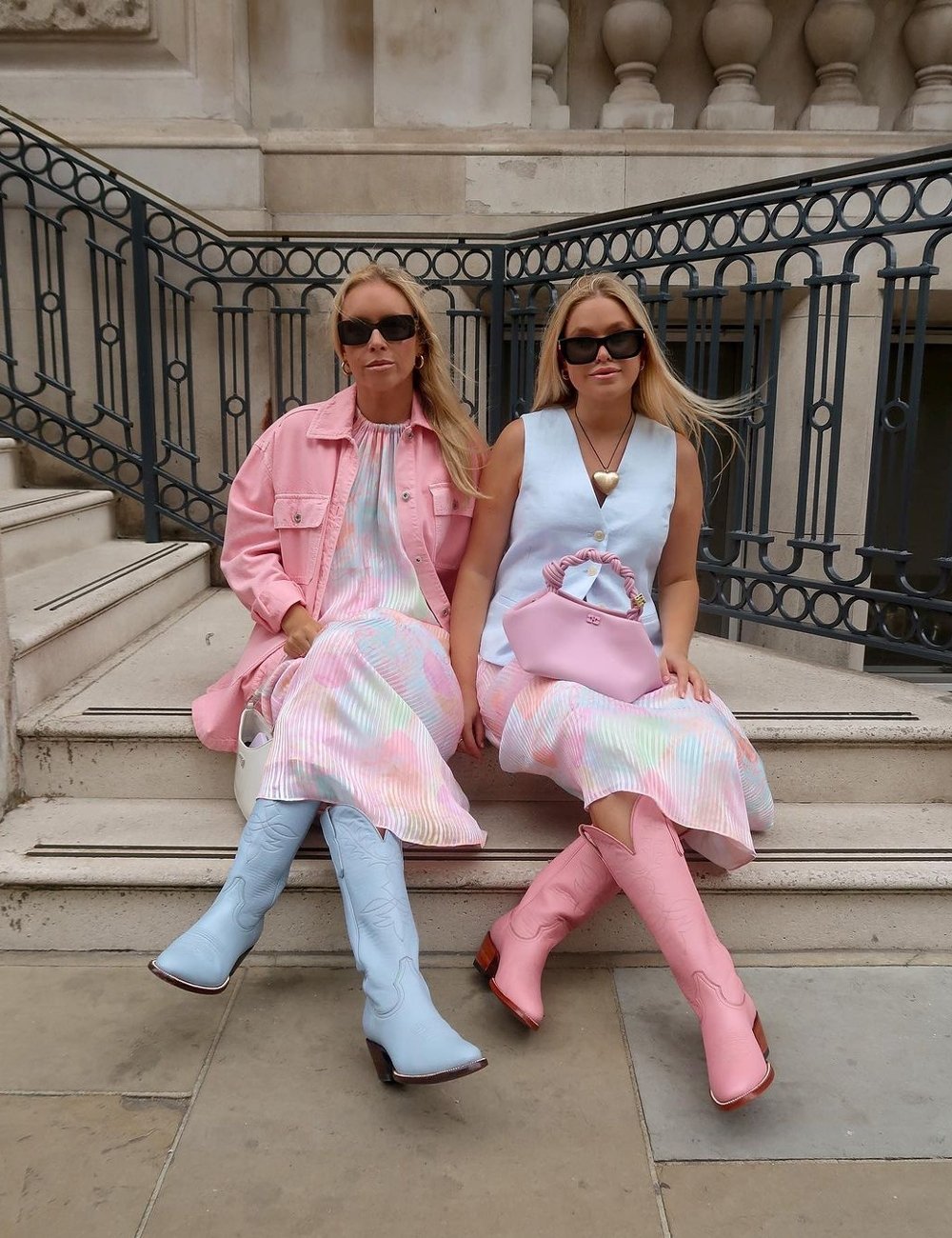 Duas mulheres sentadas usam vestidos claros e coloridos, sendo um complementado com jaqueta rosa. Ambas vestem sapatos coloridos e óculos escuros retrô. Bolsas em tons coordenados completam o look moderno e estiloso.