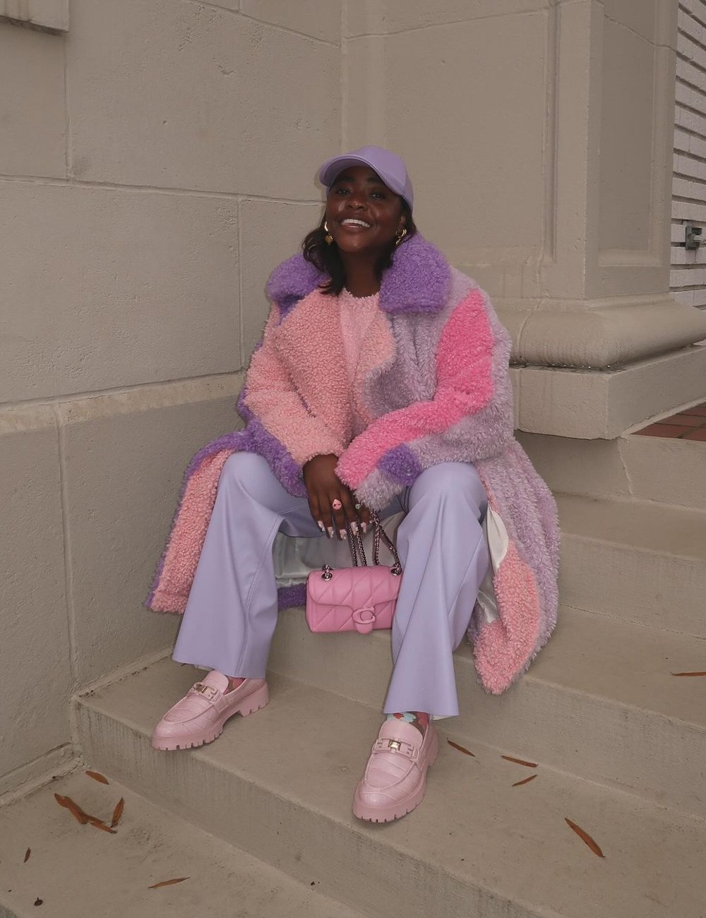 Mulher vestindo casaco oversized felpudo nas cores rosa e lilás, calça lilás, bolsa rosa e mocassins rosa. Ela usa um boné lilás e brincos dourados, destacando um visual moderno e colorido, com tons pastel. Sapatos coloridos.