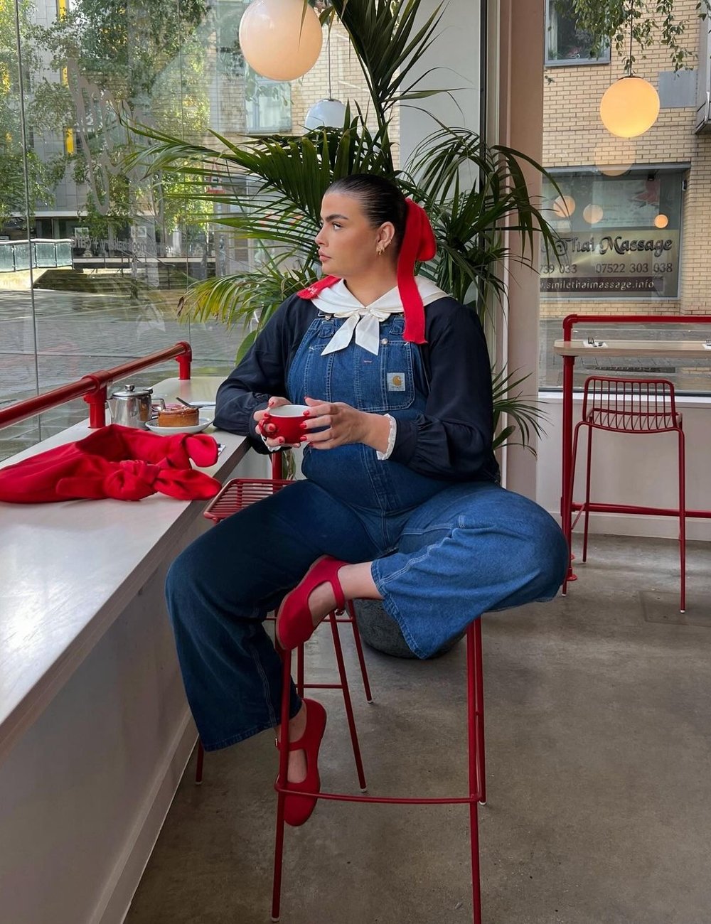 Pessoa sentada em uma cafeteria usando macacão jeans, camisa de manga longa preta com gola branca, e laço vermelho no cabelo. Sapatos coloridos vermelhos combinam com os detalhes da roupa. Look contemporâneo com toque retrô em ambiente moderno.