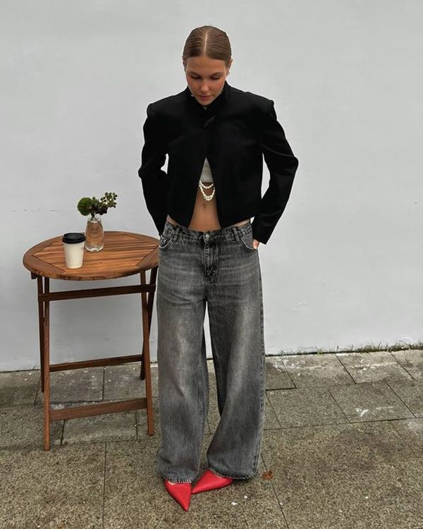 Pessoa usa tendência casual chic: blazer cropped preto, calça jeans larga de lavagem desbotada, sapatos bico fino vermelhos e top cropped com colar de pérolas. Look moderno e arrojado.