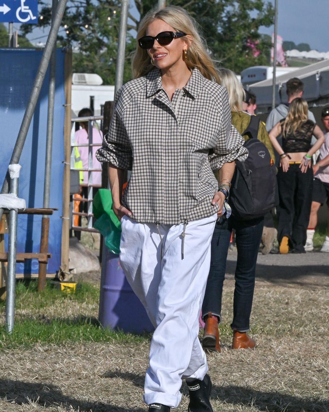 Mulher vestindo uma camisa xadrez oversized, calça branca larga com cordão ajustável na cintura, óculos de sol pretos e botas pretas. Estilo casual e despojado, típico de festivais de música. 