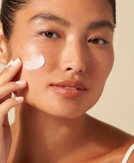 Mulher aplica creme facial em rotina de skincare. Pele saudável e maquiagem minimalista destacam tendência de beleza natural. Unhas bem cuidadas em tom nude complementam o visual. Look clean e sofisticado enfatizando auto-cuidado e bem-estar.