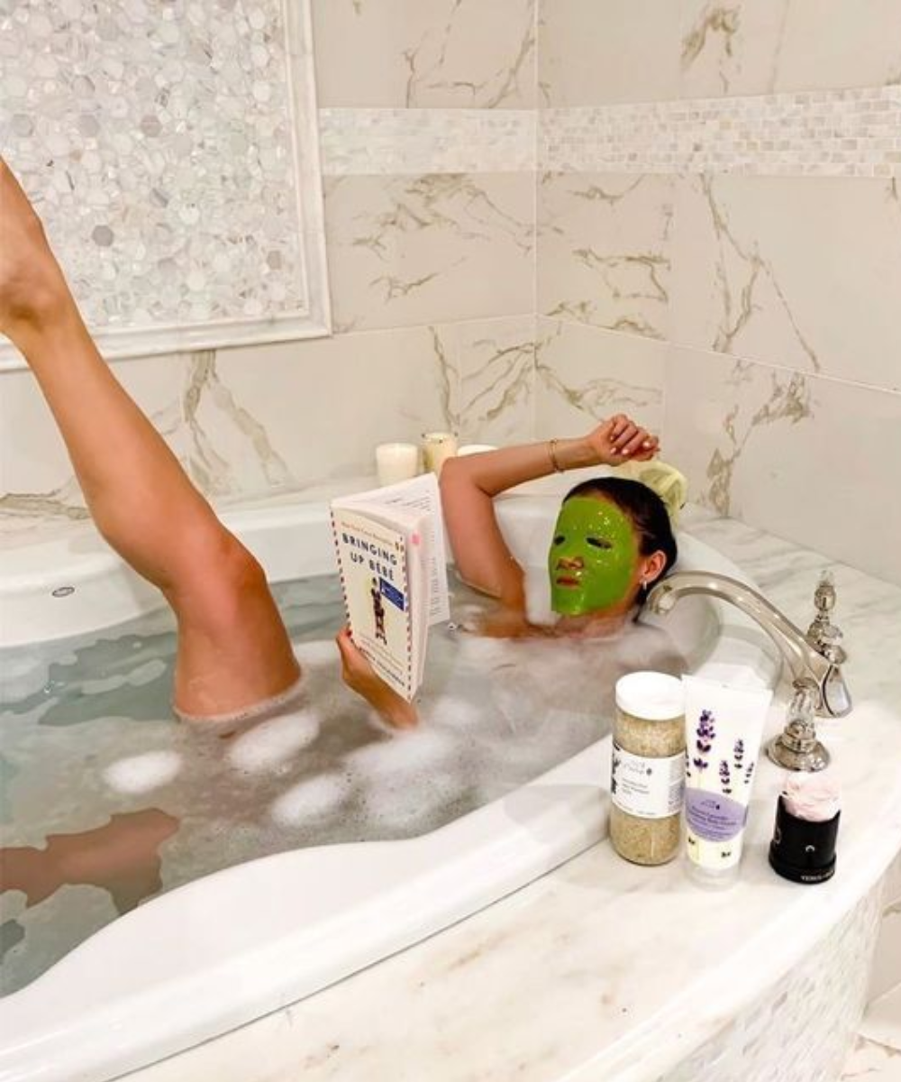 Mulher relaxa na banheira com máscara facial verde, lendo um livro. Ambiente luxuoso e cuidados com a pele indicam tendência de autocuidado e bem-estar. Produtos de beleza e detalhes em mármore ao redor da banheira denotam um estilo moderno e sofisticado.