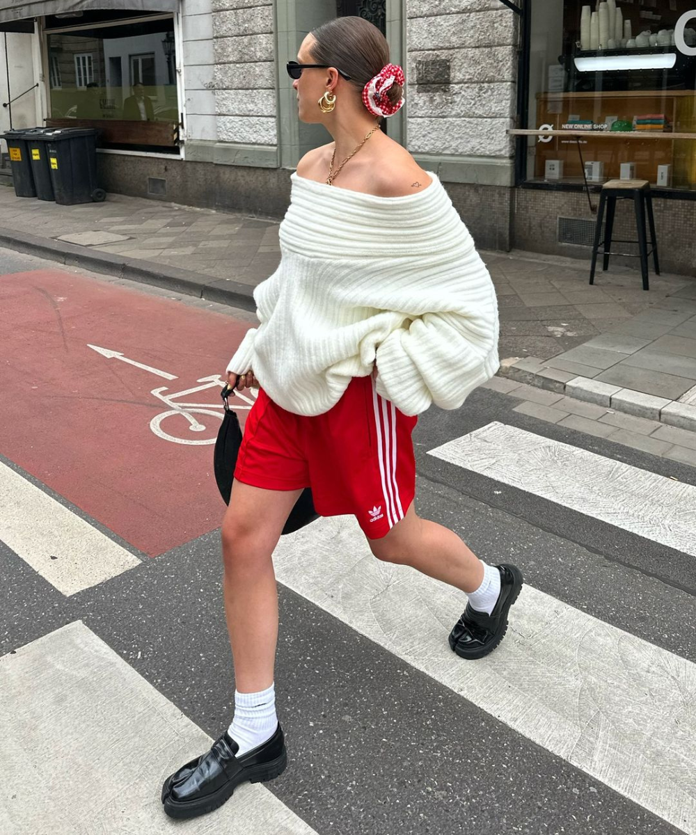 Mulher usando suéter branco oversized de tricô, shorts esportivos vermelhos Adidas, mocassins pretos com meias brancas, óculos escuros, scrunchie no coque, brincos dourados e bolsa de mão preta. Estilo casual e esportivo com toque urbano e moderno.