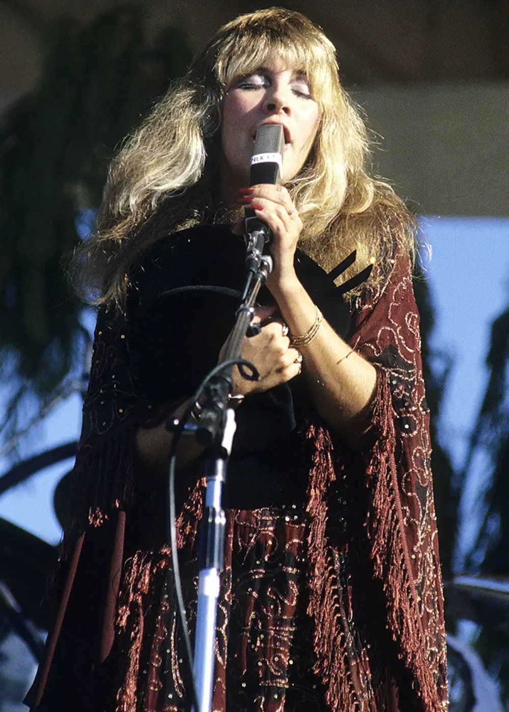 Cantora com visual boêmio, usando um vestido marrom com bordados e franjas, segurando um chapéu preto e microfone. O estilo é típico dos anos 70, com cabelos longos e ondulados, destacando tendências vintage e hippie chic da moda da época.