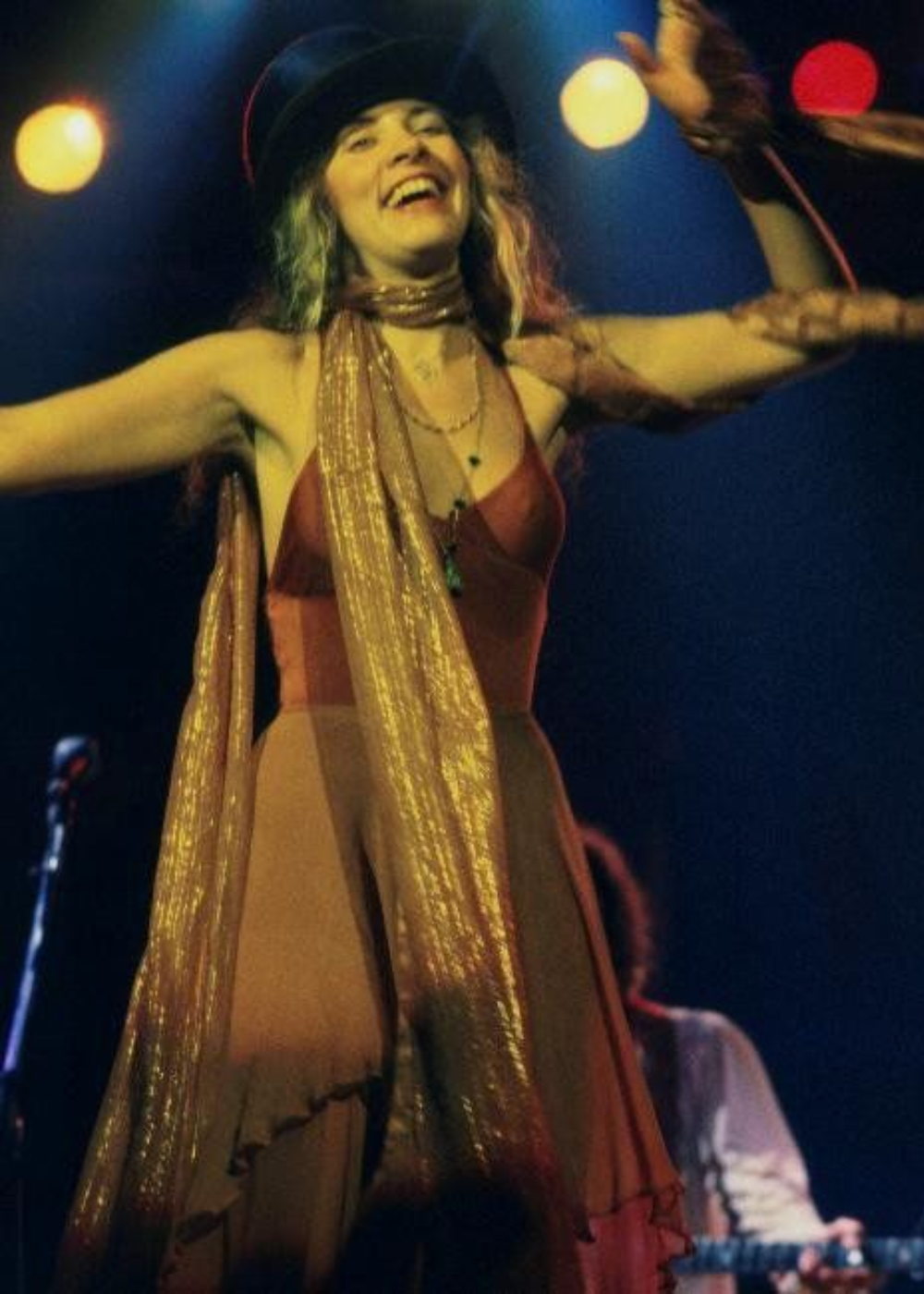 Cantora com vestido longo fluido alaranjado e chapéu preto, usando dois colares, um tecido brilhoso ao redor do pescoço e nos braços. Moda boho dos anos 70, shows ao vivo. Estilo despojado e vintage.