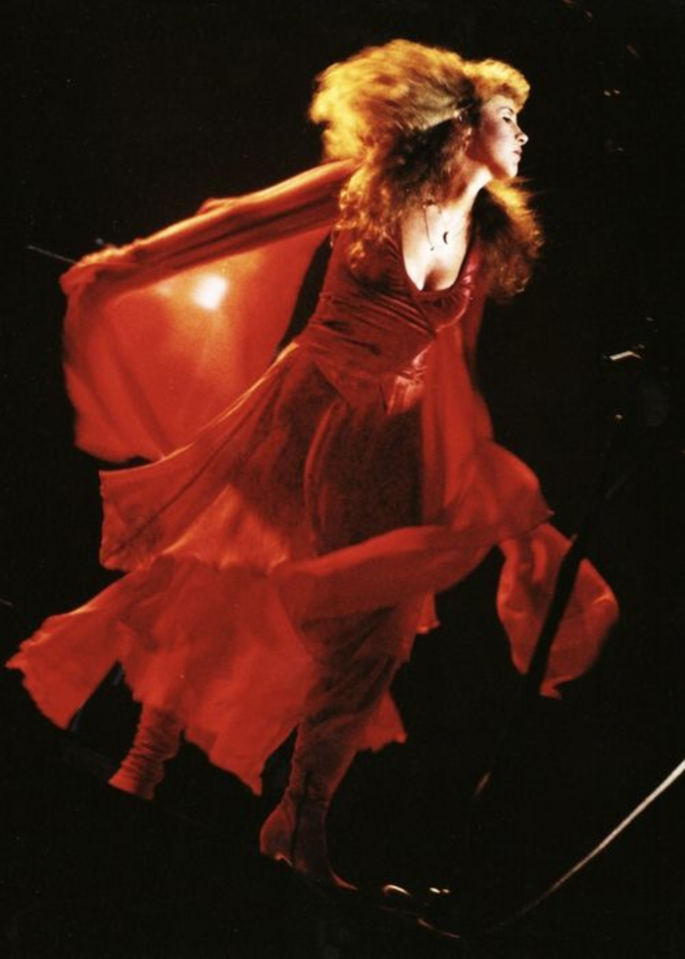 Artista com vestido vermelho longo e esvoaçante se apresenta em um palco escuro. O visual dramático, com mangas e saia fluídas, remete às tendências boho e vintage, destacando o movimento e elegância na moda dos anos 70 e 80.