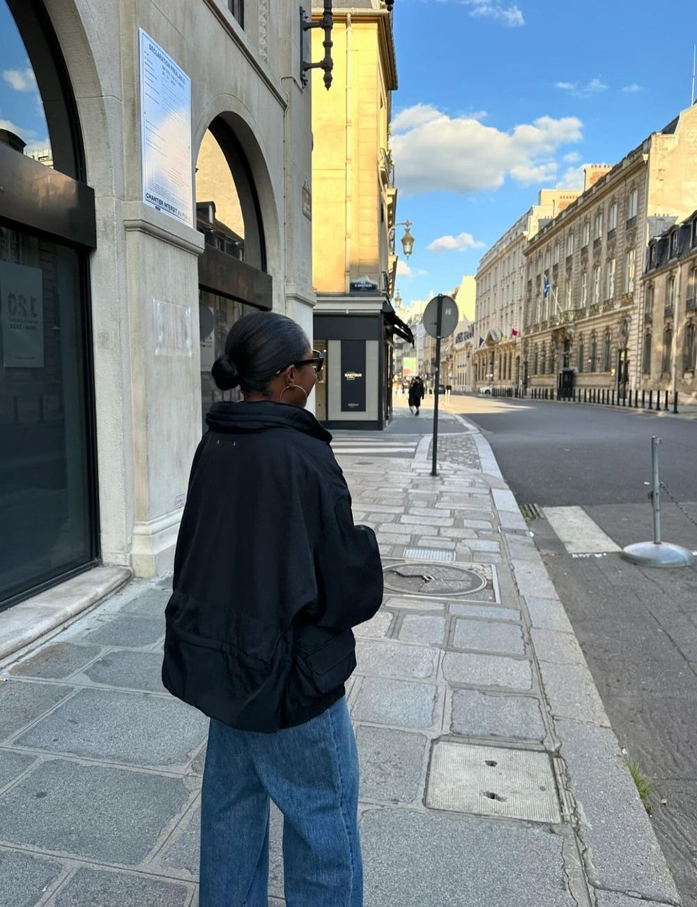 Pessoa com penteado coque baixo usa jaqueta preta oversized e jeans largos, andando em uma rua de arquitetura clássica. O look casual inclui óculos de sol escuros, refletindo tendência de moda urbana e conforto.