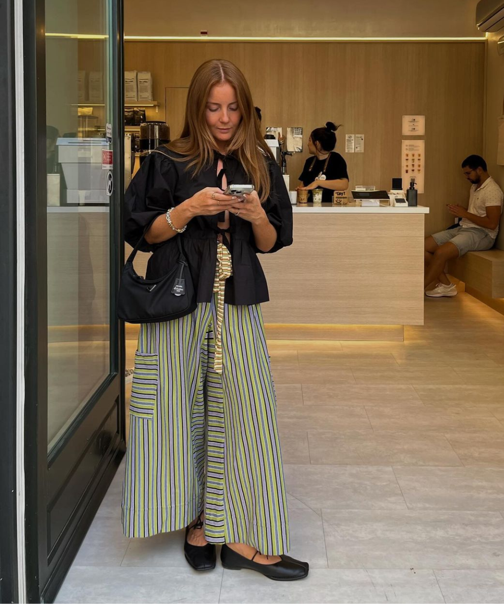 Mulher de cabelo comprido usando blusa preta com mangas bufantes e calça larga listrada nas cores amarelo, azul e cinza. Acessórios: bolsa preta de ombro e sapatilhas pretas. Moda casual e confortável, combinando peças amplas e cores neutras.