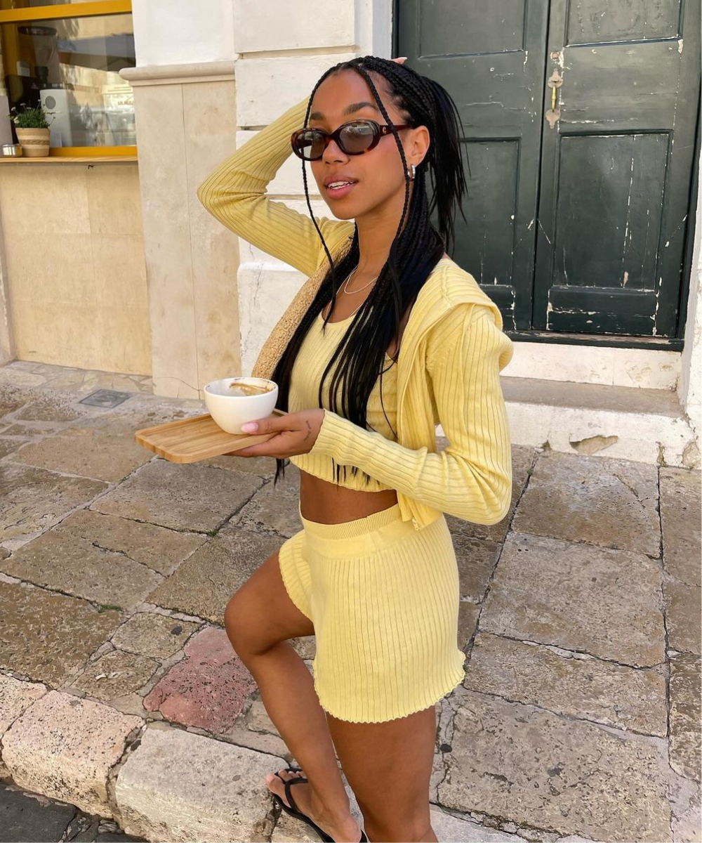 Mulher com cabelo trançado e óculos de sol usa conjunto de tricô amarelo, combinando top cropped, shorts e cardigan. Ela segura uma xícara de café em uma bandeja de madeira. Look casual e confortável, ideal para dias ensolarados.