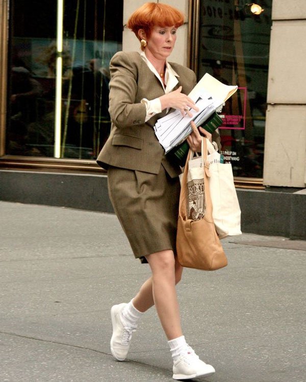 Mulher vestindo um conjunto de terninho verde-oliva com saia na altura dos joelhos, camisa branca e tênis brancos com meia. Carrega bolsas e papéis, conferindo um estilo profissional, porém prático e confortável para o dia a dia urbano.