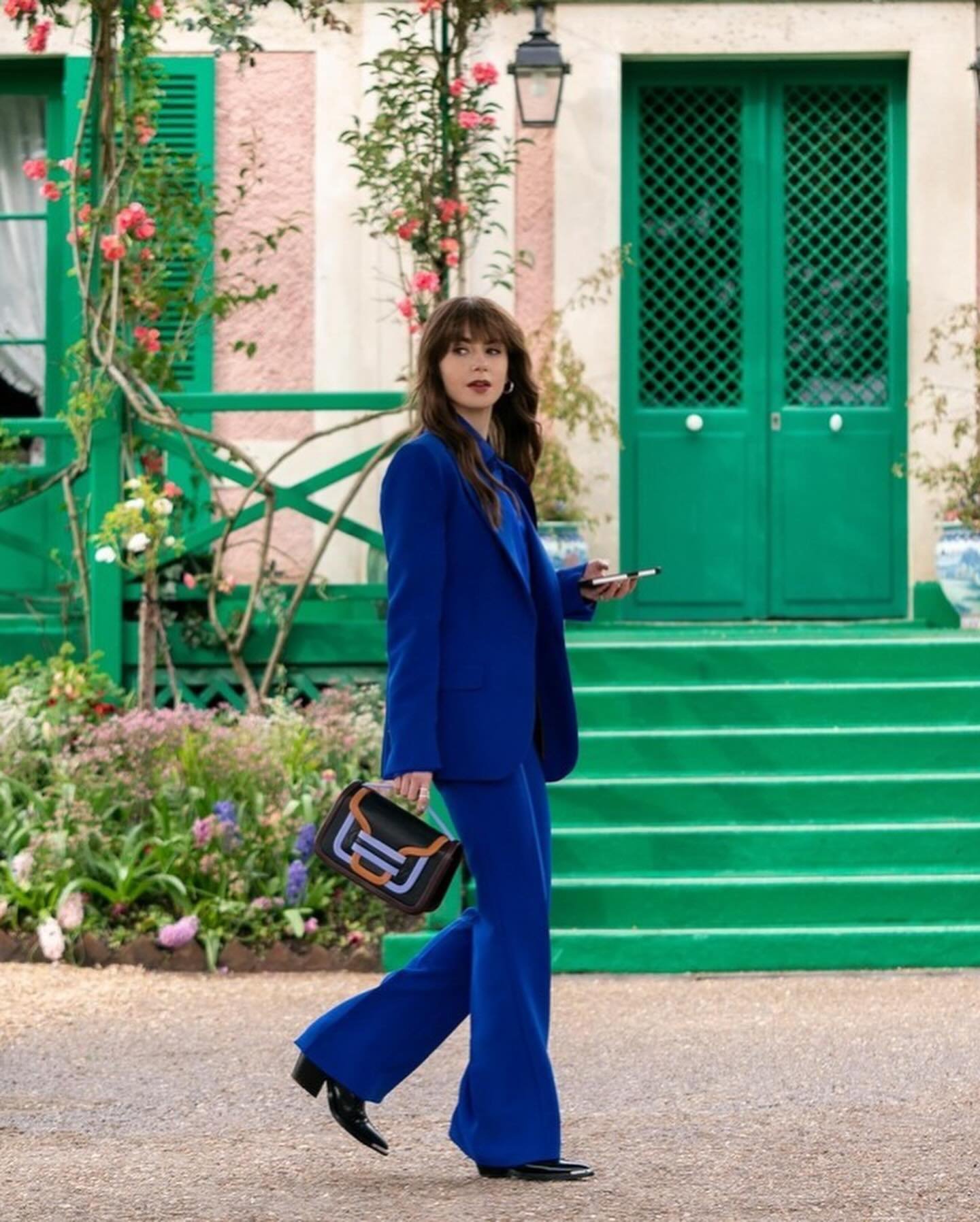 Mulher usando um elegante terninho azul royal em corte reto, tendência de alfaiataria moderna. Ela carrega uma bolsa de ombro estilosa preta com detalhes coloridos, combinando com sapatos clássicos pretos. Ambiente externo com fundo de verde e flores.